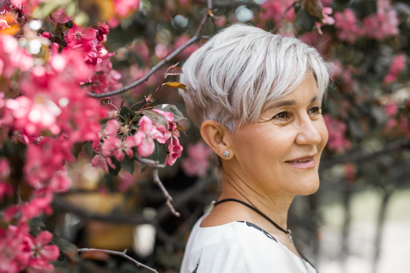 Smiling woman with short silver hair standing beside pink flowering branches outdoors, looking to the side in soft natural light.