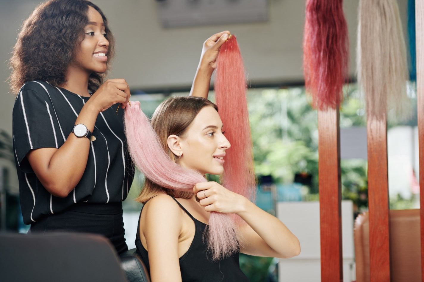 A hairstylist helping a woman compare pink-toned hair extensions during a custom wig or hair extension consultation in a salon setting.