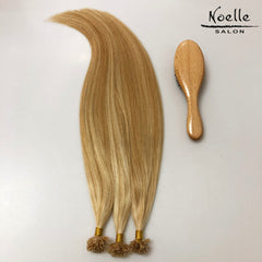 Three bundles of blonde hair extension tips with gold bands are displayed next to a wooden paddle brush on a plain white background, with the 'Noelle SALON' logo in the top right corner.